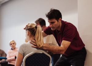 A physio examines the neck motion of a person with a ponytail sitting a chair.