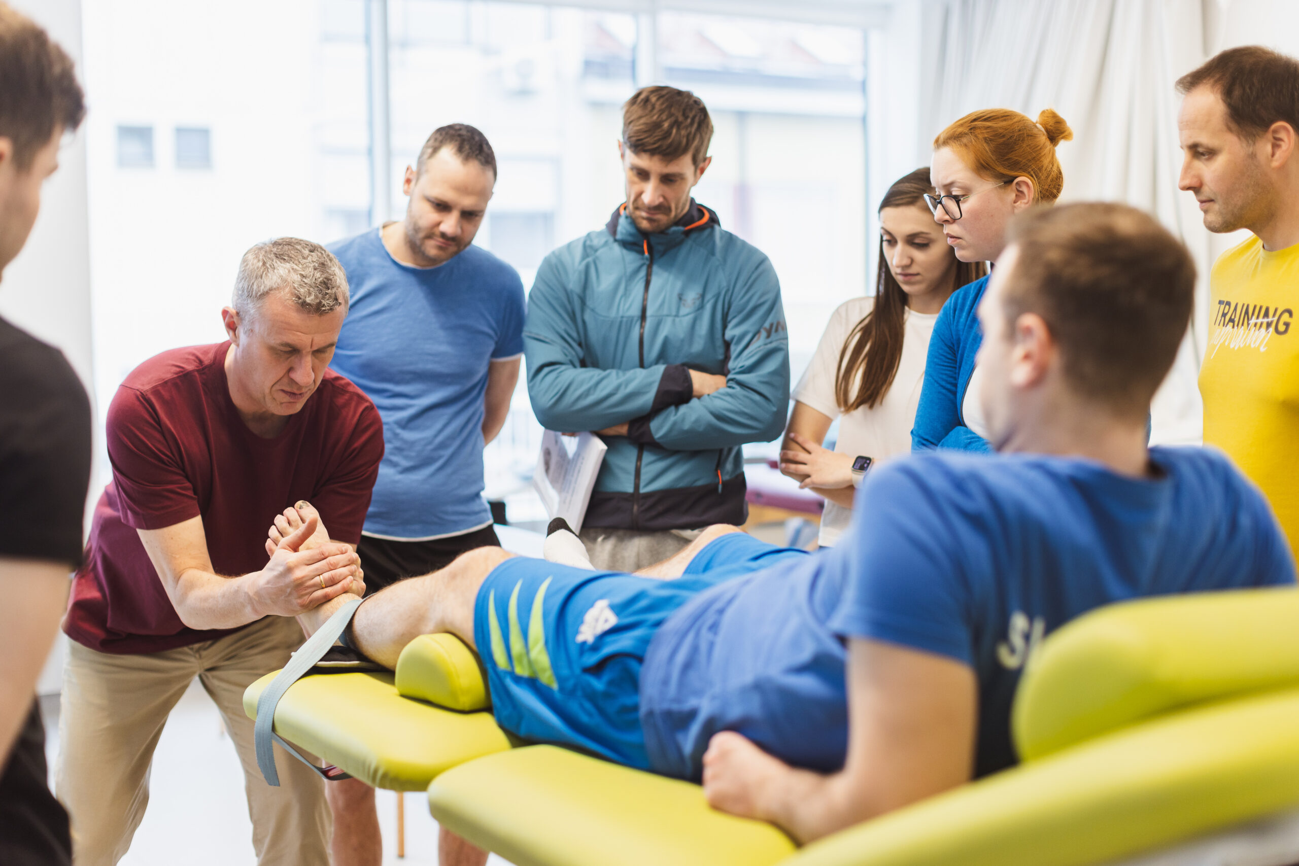 Seven physios watch while a KEOMT instructor teaches them how to perform an ankle mobilization.